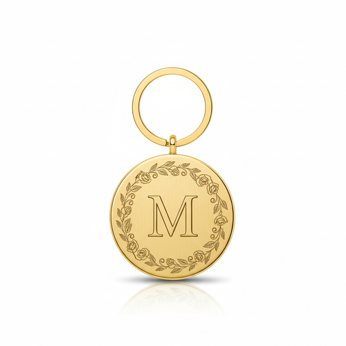 Gold metal disc keychain with letter M in elegant serif font surrounded by delicate floral engraving on white background