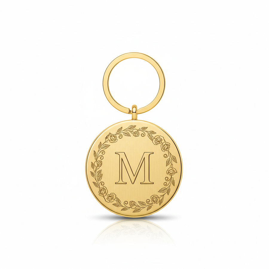 Gold metal disc keychain with letter M in elegant serif font surrounded by delicate floral engraving on white background